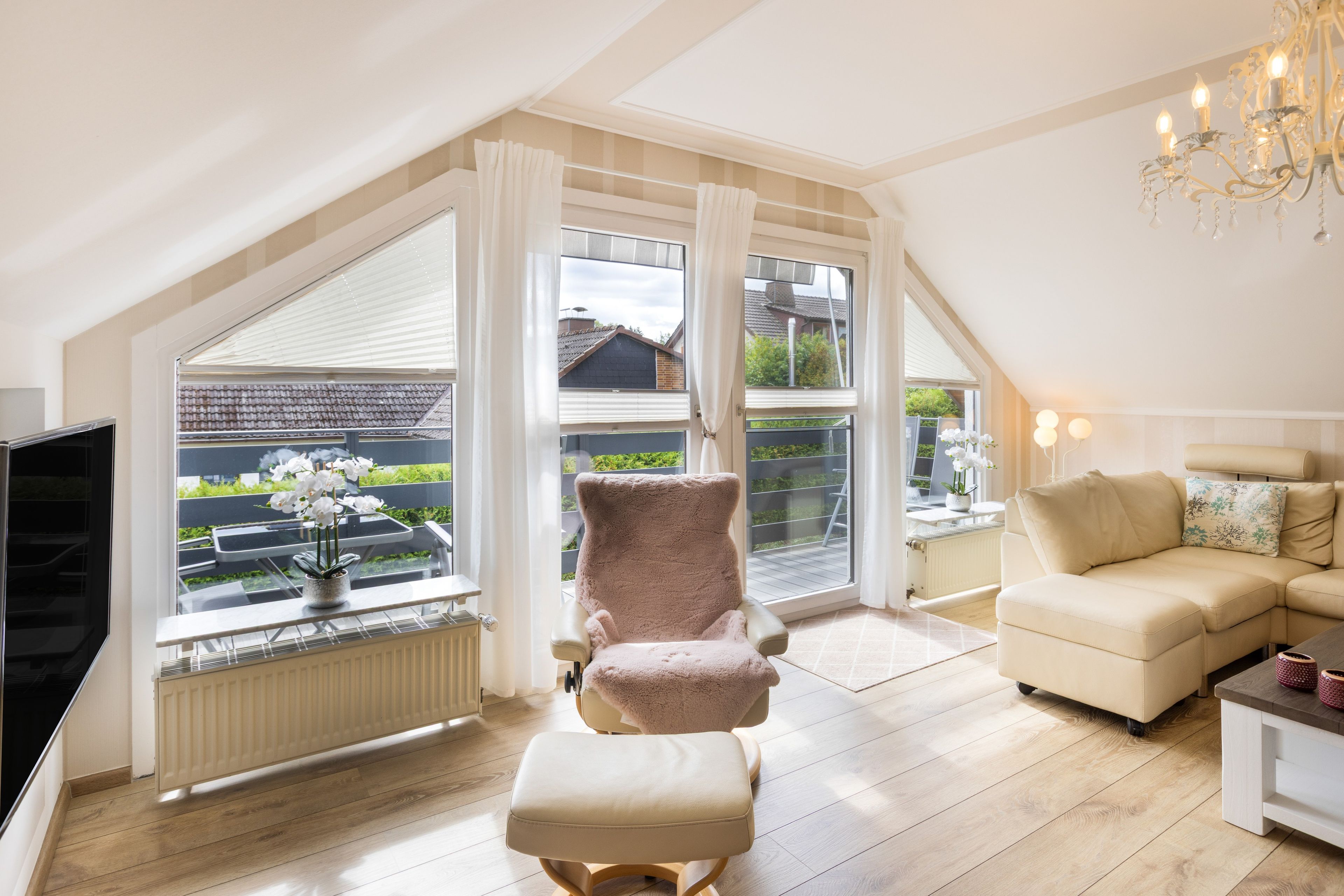A spacious living room with a high ceiling, large windows, a comfortable sofa, a plush armchair, and a television.