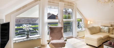 A spacious living room with a high ceiling, large windows, a comfortable sofa, a plush armchair, and a television.