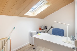 Una habitación con techo inclinado, tragaluces, radiador, escritorio, silla, ventilador y aspiradora.