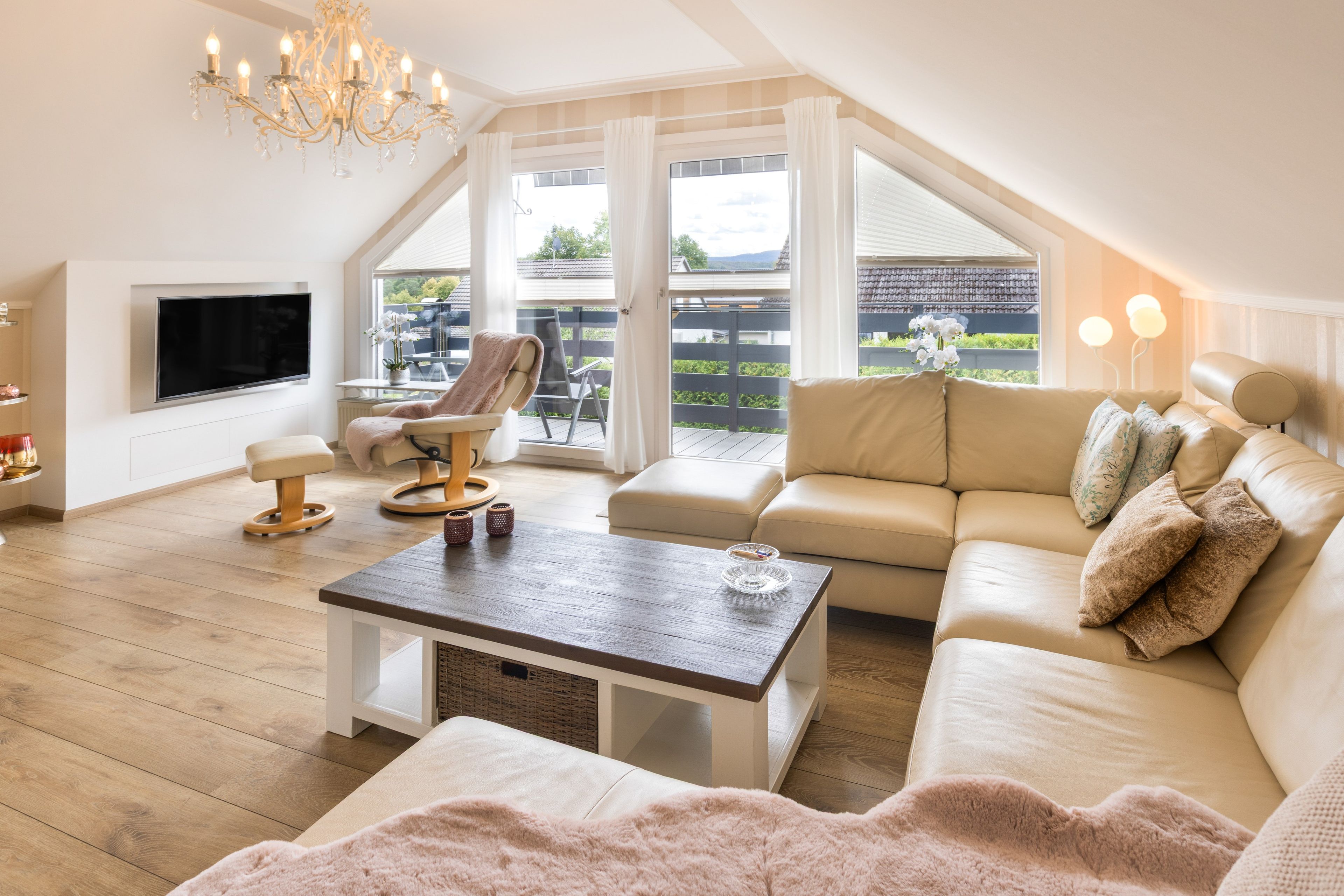 A spacious living room with a high ceiling, a flat-screen TV mounted on the wall, a comfortable sofa set, a coffee table, and a view of the outdoors through large windows.