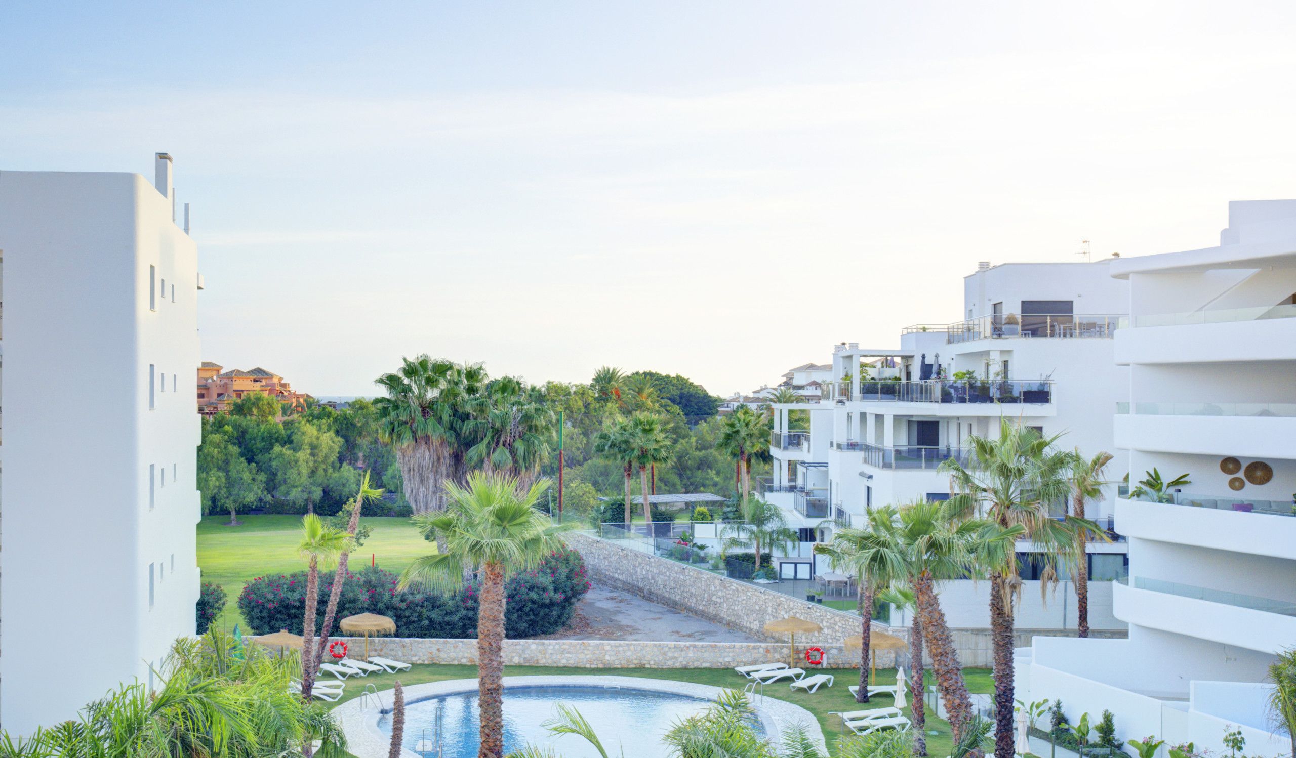 Ein Resort mit Pool, umgeben von Palmen und Liegestühlen, moderne Architektur und Blick auf weitere Gebäude im Hintergrund.