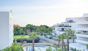 Un resort con piscina rodeada de palmeras y sillones de descanso, arquitectura moderna y vistas a otros edificios al fondo.