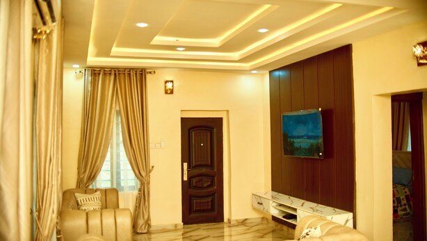 A modern living room with a sofa, a television mounted on the wall, and a door with a decorative panel. - Pinefield Courts Apartmentl  (Akure)