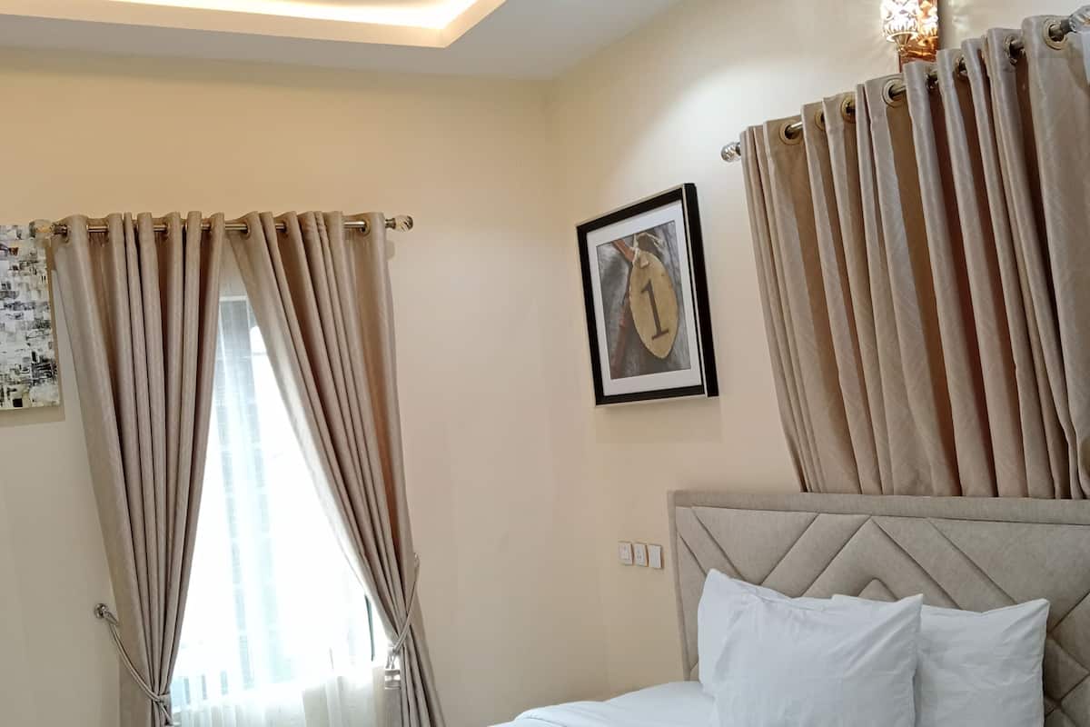 A hotel room with a bed, beige curtains, a framed picture on the wall, and recessed ceiling lighting.