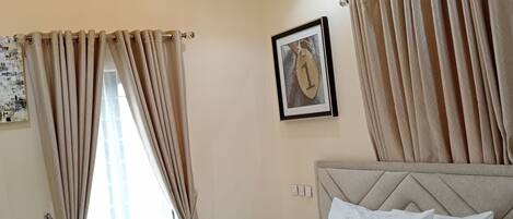 A hotel room with a bed, beige curtains, a framed picture on the wall, and recessed ceiling lighting.