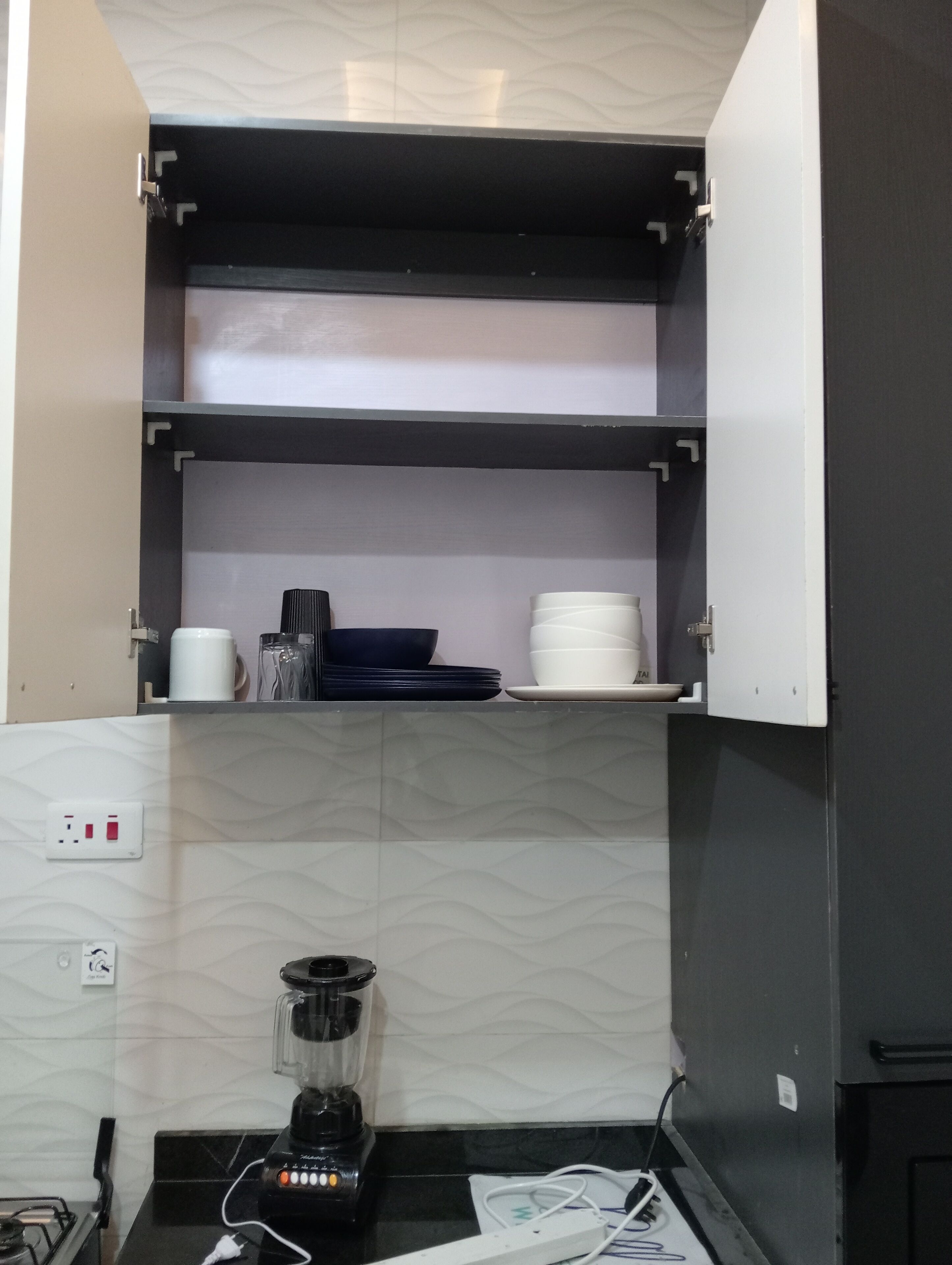Una cocina con una licuadora empotrada en la encimera, una estufa a gas y un microondas. Hay dos estantes encima de la encimera, uno con platos y tazas, y otro con un recipiente negro y un bol blanco.