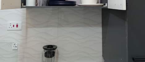 Una cocina con una licuadora empotrada en la encimera, una estufa a gas y un microondas. Hay dos estantes encima de la encimera, uno con platos y tazas, y otro con un recipiente negro y un bol blanco.