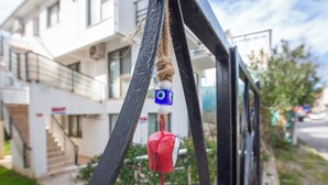 A red and white bell hanging from a black metal structure, with a blue cube with an eye symbol attached to it. - Poseidon Kinaliada Pansiyon (Adalar)