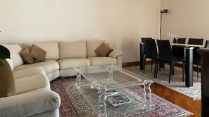 A living room with a sofa, a glass coffee table, a dining table with chairs, and a patterned rug.
