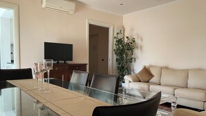 A modern living room with a glass dining table, leather chairs, a beige sofa, and a television.