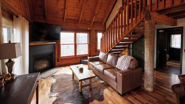 A cozy cabin interior with a fireplace, leather sofa, wooden floors, and a staircase.