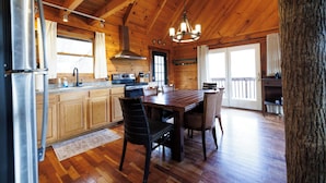 A wooden cabin interior with a dining table, chairs, a kitchen area, and a large window.