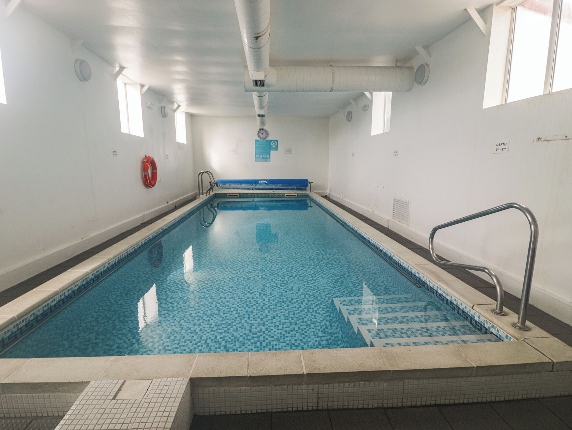 An indoor swimming pool with clear blue water, a lifebuoy, and a depth marker.