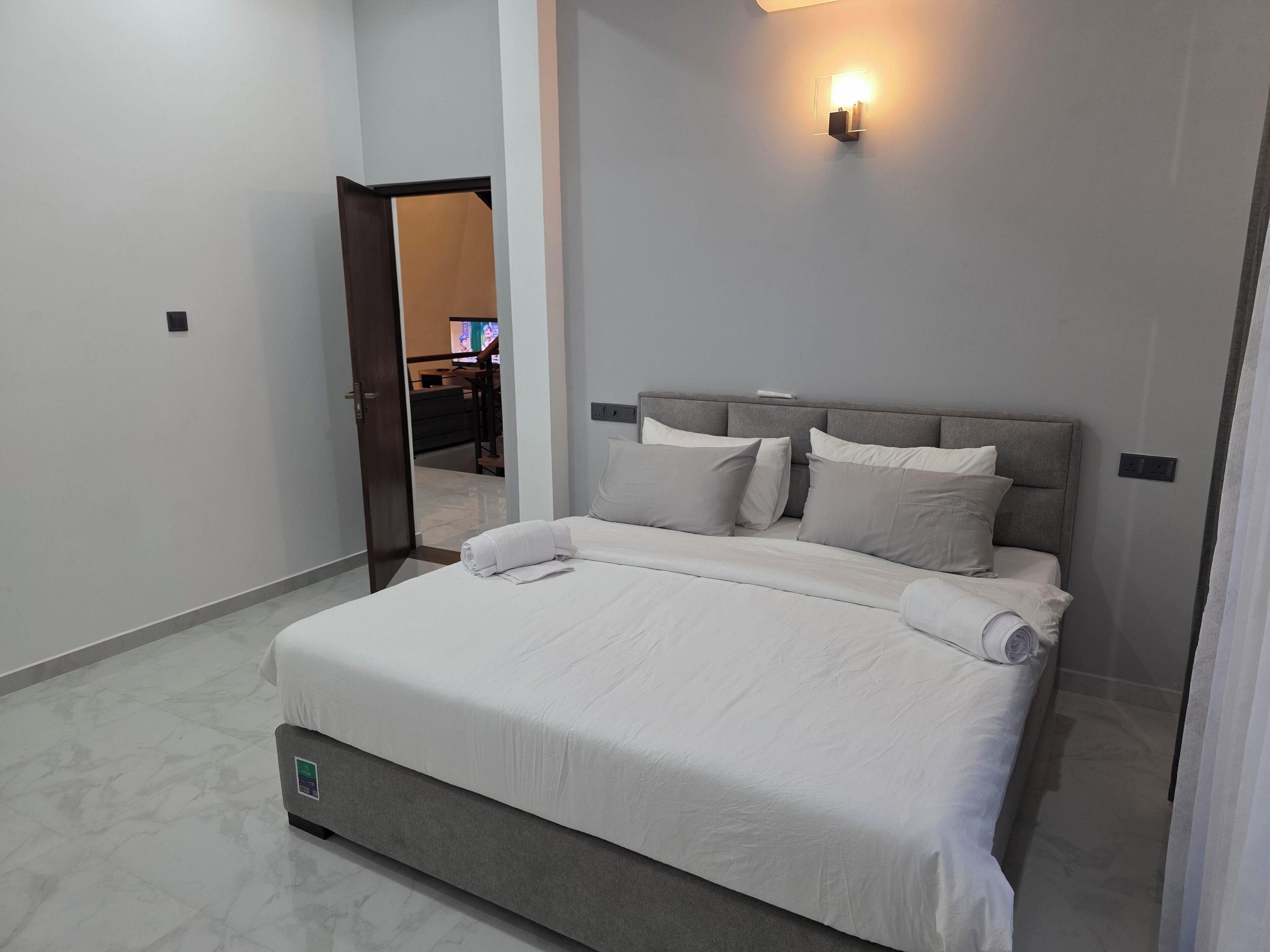 A neatly made bed with white linens and pillows, a grey headboard, and a wall-mounted light fixture.