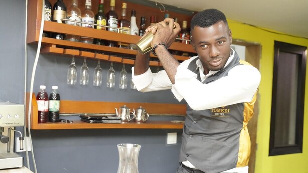 En bartender bag baren med hylder fyldt med flasker og glas, en kaffemaskine og et vinglas.
