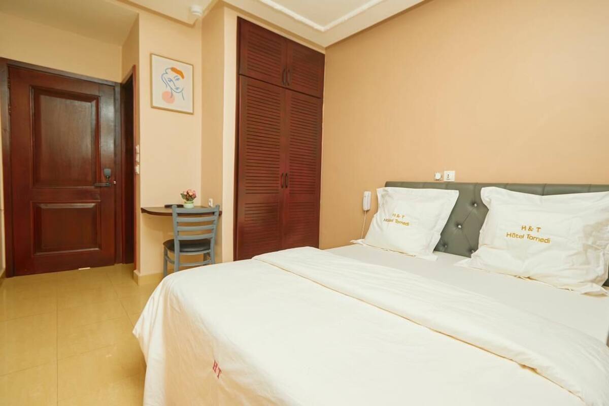 A hotel room with a bed, a desk, a chair, a wardrobe, and a wall-mounted telephone.