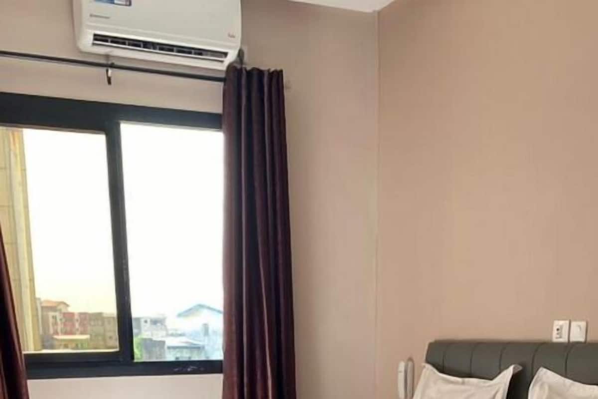 A hotel room with a bed, a chair, a window with curtains, and an air conditioning unit.