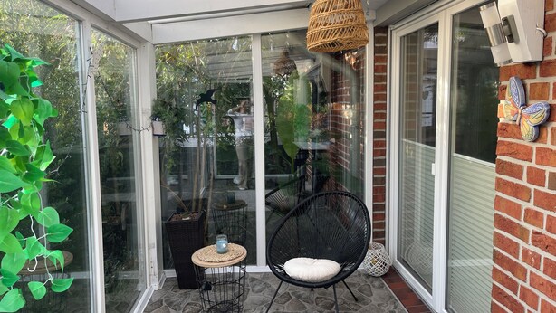 Un patio cubierto con puertas de cristal, un colgante de luz de mimbre, una silla negra con cojín y una mesita redonda.