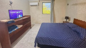 Un dormitori amb un llit, una televisió sobre un suport de fusta, aire condicionat i una finestra amb cortines.