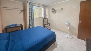 Un dormitori amb un llit, una cadira, un escriptori, una finestra amb cortines, una porta i dos ventiladors de paret.