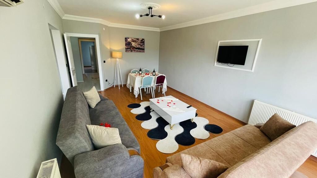 Una sala de estar moderna con un sofá gris, un sofá beige, una mesa de centro y una mesa de comedor con sillas. Hay un televisor montado en la pared y un radiador en el suelo.
