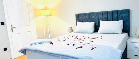 A neatly made bed with a blue headboard, white pillows, and a light blue bedspread. There are rose petals scattered on the bed.