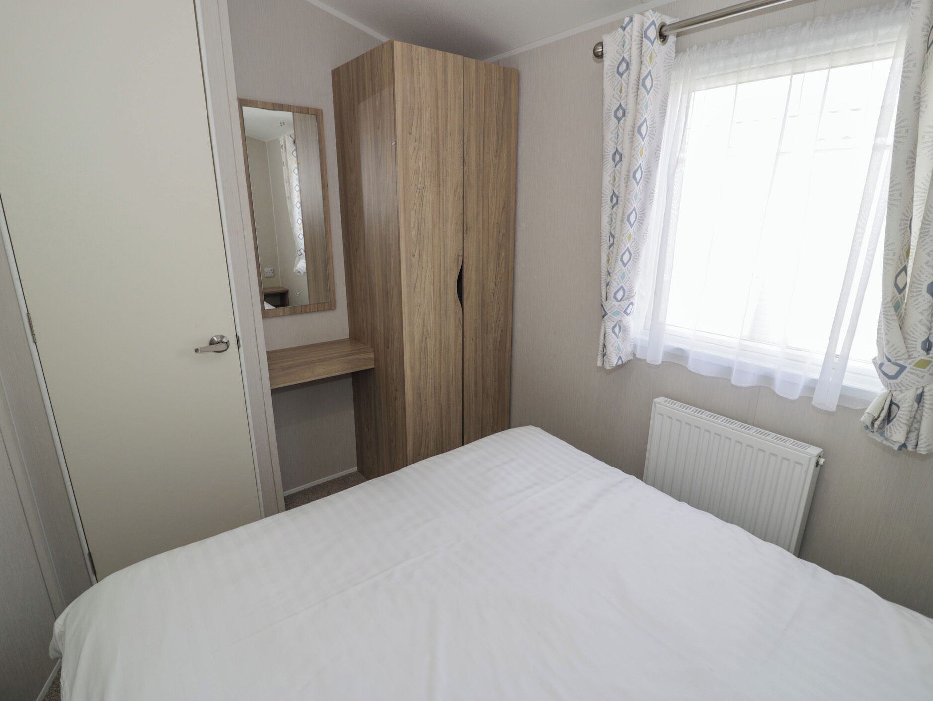 Un dormitorio con cama, armario, espejo y ventana con cortinas.