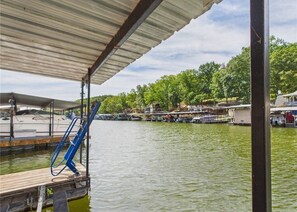 Un muelle con una zona cubierta, rampa para embarcaciones y vistas a un lago con barcos y edificios al fondo.