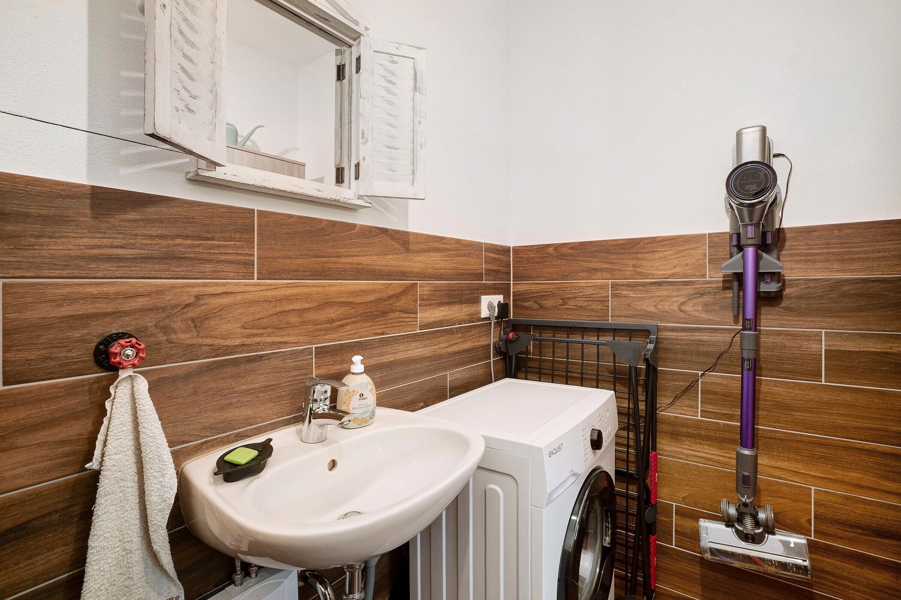 Un bagno con lavandino, specchio, asciugamano, lavatrice e aspirapolvere.