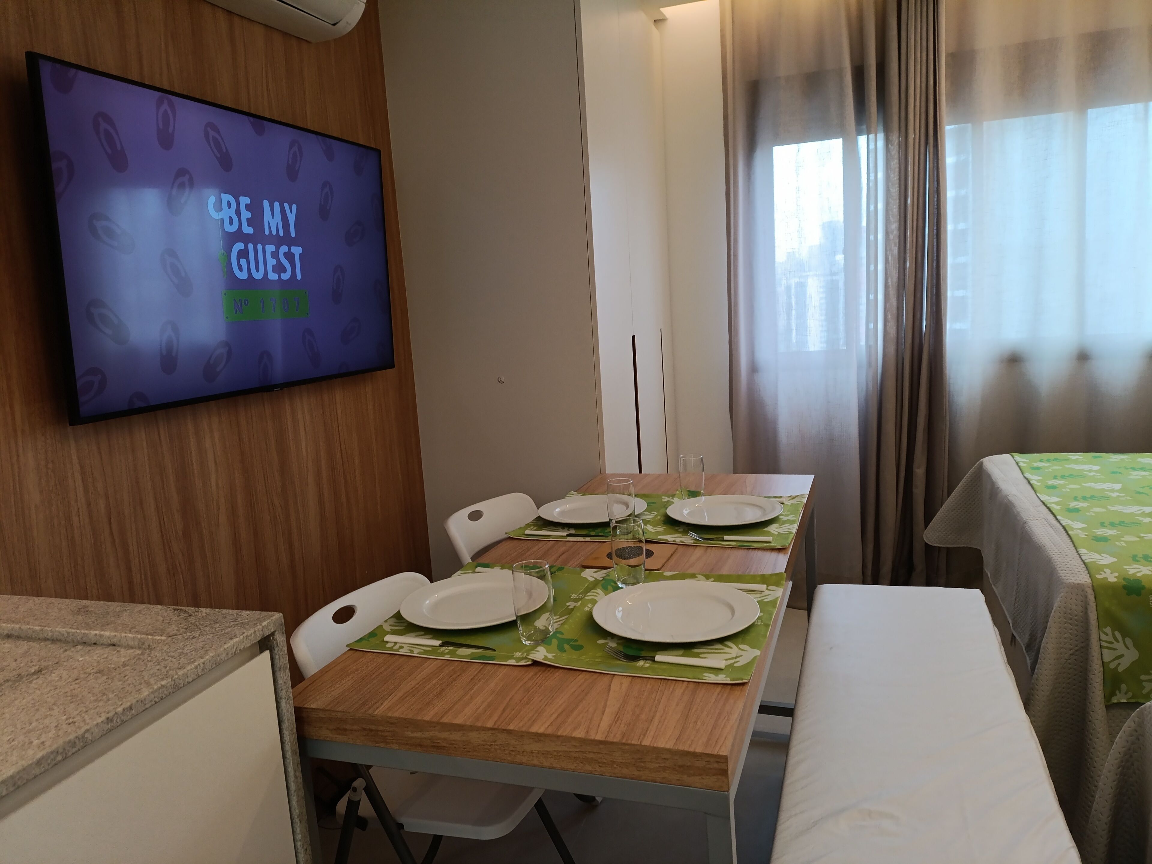 Ein Hotelzimmer mit Esstisch für vier Personen, einem Fernseher, der "BE MY GUEST" anzeigt, und einem Fenster mit Vorhängen.