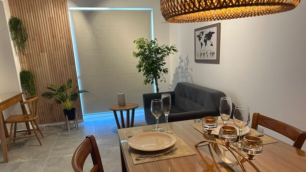 Un coin repas avec une table en bois dressée pour six personnes, entourée de chaises en bois. On y trouve également une petite table ronde avec un vase et une plante en pot. Un canapé gris est visible au fond.