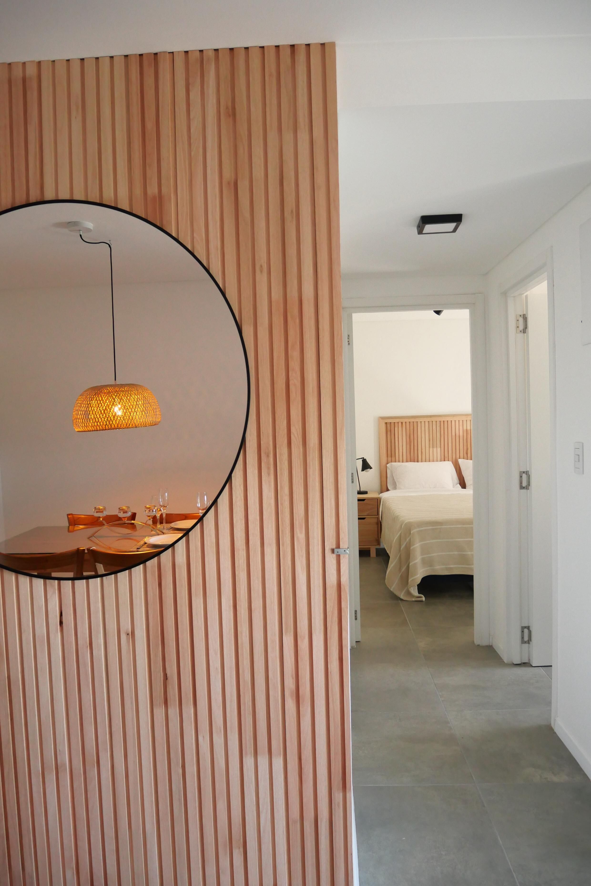 Une chambre moderne avec un mur lambrissé en bois, un grand miroir rond, un luminaire suspendu, un lit soigneusement fait et une porte visible donnant sur une autre pièce.