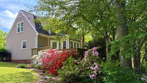 Una casa de dos pisos con un porche, rodeada de abundante vegetación y plantas con flores.