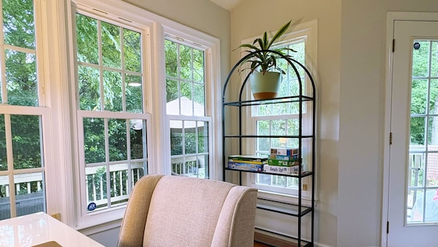 Una habitación con una mesa blanca, una silla color beige, un estante metálico negro con una planta y ventanas amplias.