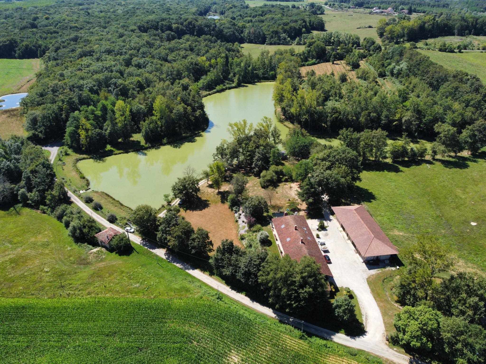 Vue aérienne d’une propriété rurale avec un étang, une végétation luxuriante et quelques bâtiments aux toits rouges.