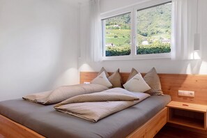 A bed with a headboard, a bedside table, a window with a view of greenery, and a power outlet on the wall.