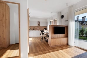 A modern kitchen with wooden cabinets, a dining area with a table and chairs, a flat-screen TV, and a large sliding glass door leading to a balcony with a view of buildings.
