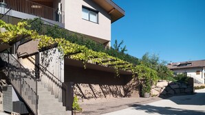A modern two-story house with a stone staircase, a balcony with climbing plants, and a clear blue sky.