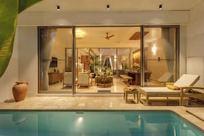 A modern indoor pool area with a glass-enclosed living room, lounge chairs, and a dining area visible through the glass.