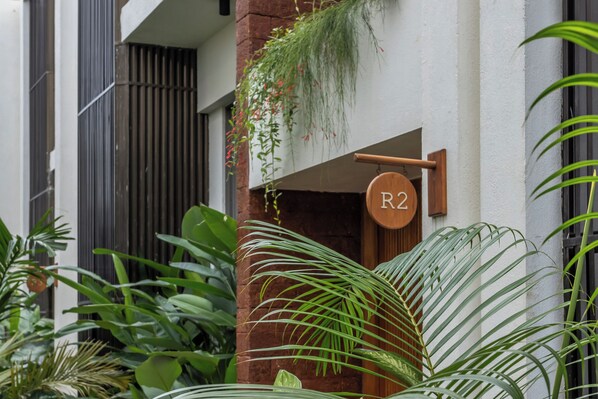 A building entrance with a wooden sign indicating room R2, surrounded by lush green plants.