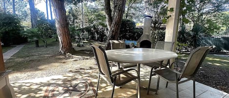 Une terrasse extérieure avec une table ronde et des chaises, entourée d’arbres et agrémentée d’une cheminée en pierre.