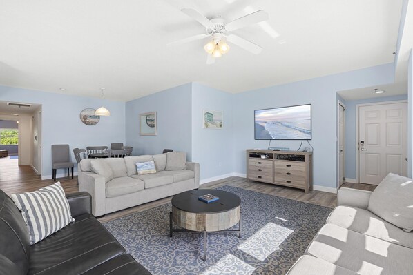 A living room with a beige sofa, a black leather couch, a round coffee table, a TV, and a ceiling fan. - Oceanwalk 4-201 (New Smyrna Beach)