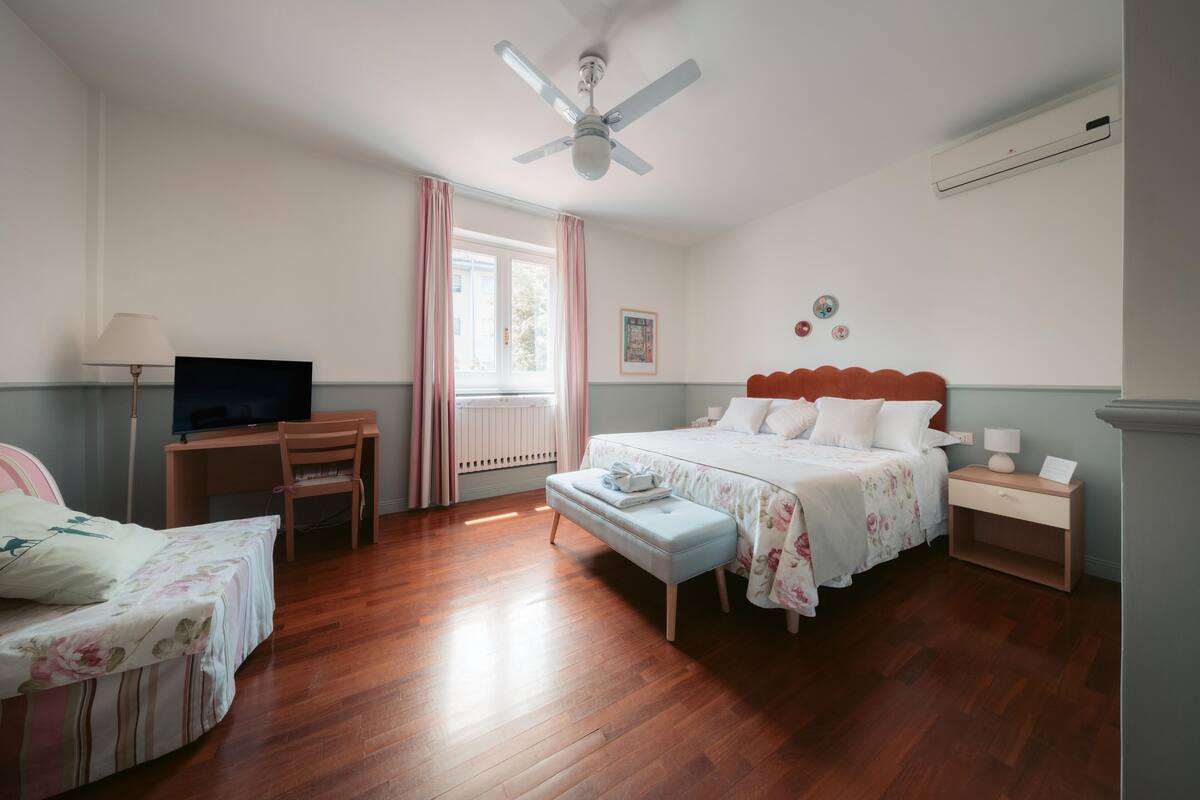 A bedroom with a bed, a desk with a chair, a television, and a ceiling fan.