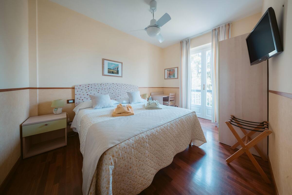 A bedroom with a bed, nightstand, bedside table, lamp, and a TV mounted on the wall.