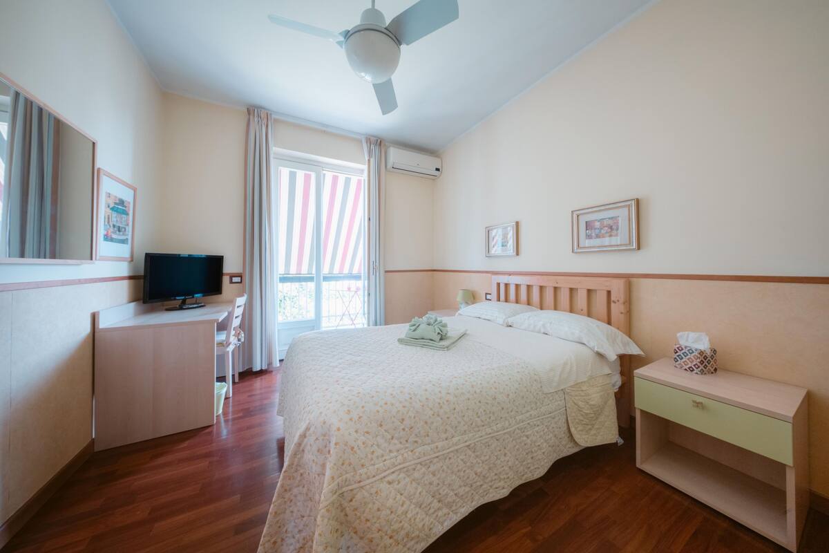 A hotel room with a bed, a desk with a television, a nightstand, and a window with curtains.