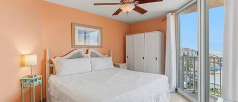 A bedroom with a bed, a ceiling fan, a nightstand, a lamp, a wardrobe, and a large window with a view.