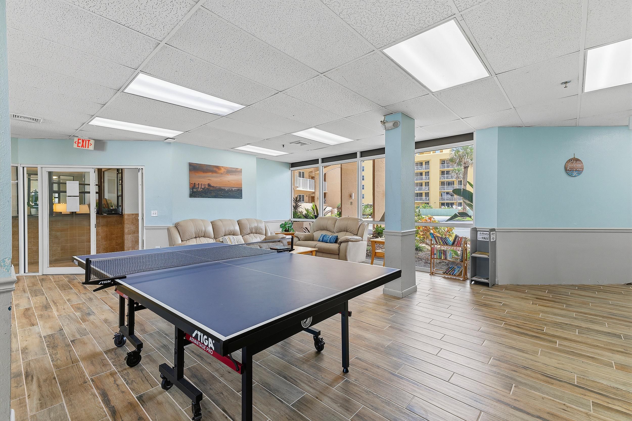 A room with a ping pong table, a sofa, a bookshelf, and a view of a building outside.