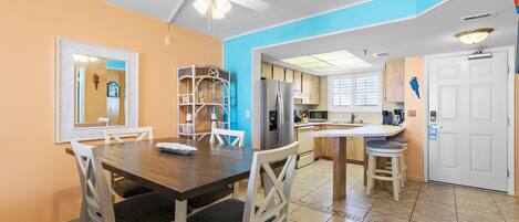 A dining area with a wooden table and chairs, a ceiling fan, and a kitchen area with cabinets and a refrigerator.