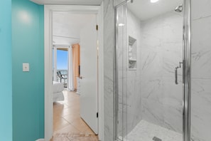 A bathroom with a glass shower enclosure, a white door, and a view of a balcony. - Oceania Plaza 808 (New Smyrna Beach)