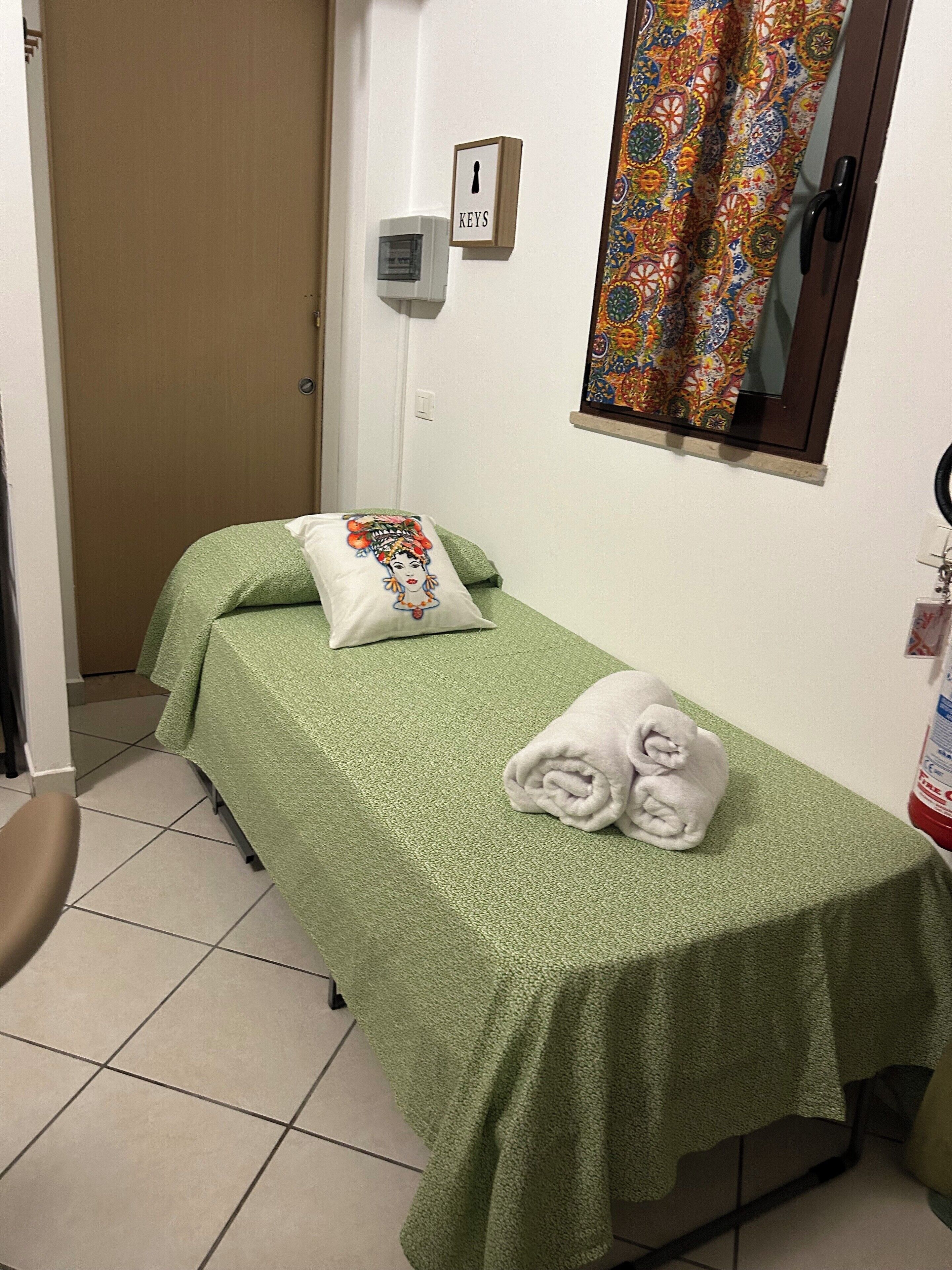 A small, single-bed room with a green bedspread, a pillow with a floral design, and a towel folded on the bed. There is a door to the left and a window with a colorful curtain to the right.